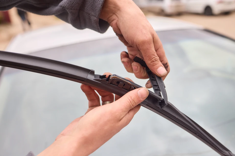 Personne installant un essuie-glace neuf sur le pare-brise d'un véhicule, manipulation des clips de fixation avec les deux mains, opération de maintenance automobile en extérieur