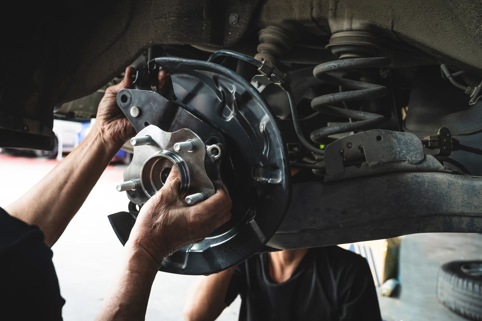 Professionnel expérimenté en tenue professionnelle dans un atelier ou un espace de stockage, examinant attentivement des pièces détachées automobiles variées (moteur, filtres, courroies, plaquettes de frein, alternateurs, etc.) organisées sur des étagères ou un établi. L'atmosphère doit être celle d'un expert qualifié maîtrisant sa sélection de pièces neuves de qualité, loin de l'atmosphère commerciale de concession automobile. Éclairage professionnel, environnement technique et organisé, reflétant 35 ans de maîtrise du métier de distribueur de pièces.